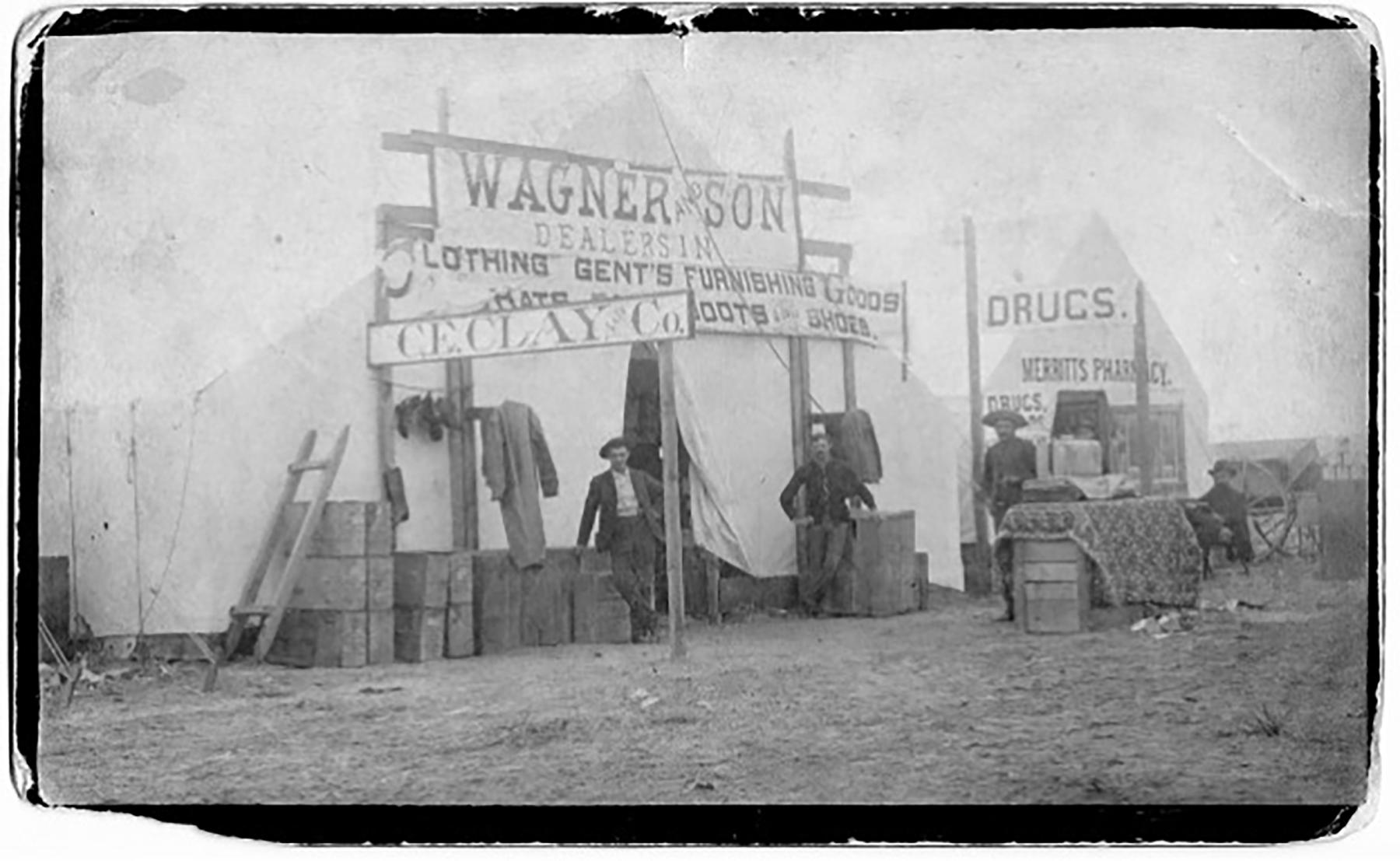 Large Canvas tent with signs: "Wagner and Son: Clothing, Gent's Furnishings, Goods, Hats, Boots, Shoes," "C.E.Clay & Co.," "Drugs, Merritt's Pharmacy." Four men pose in front, along piles of wood boxes