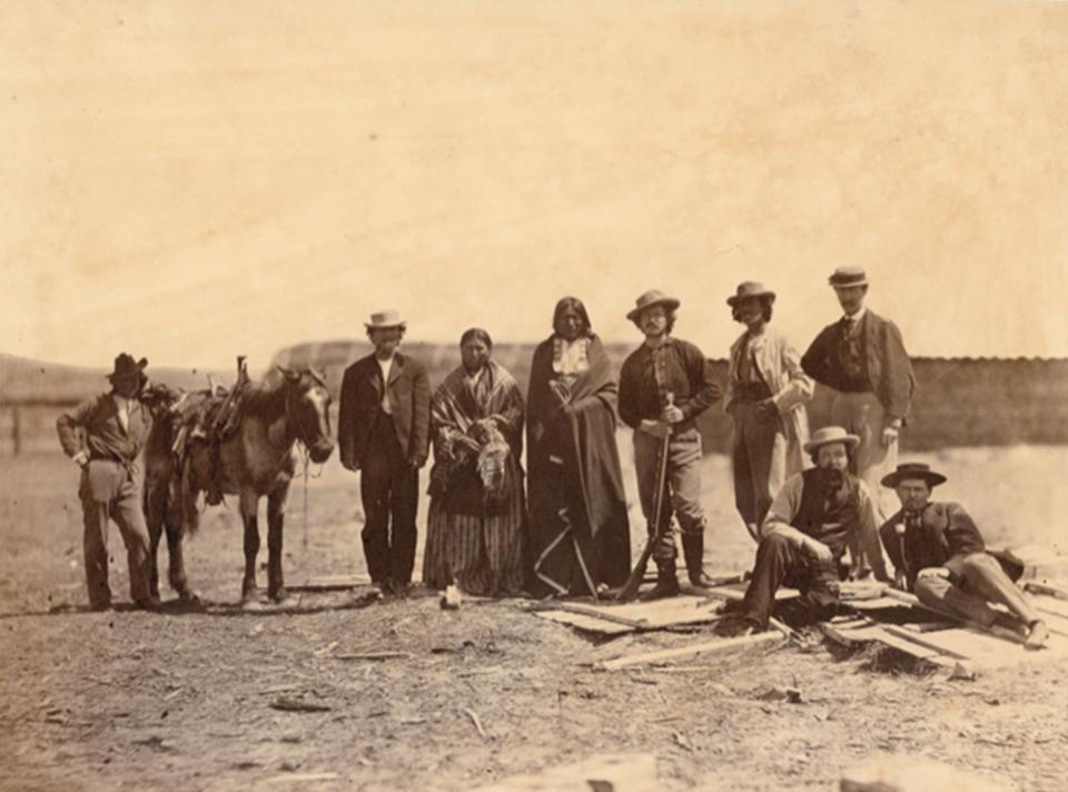 Lakota Sioux men and non-Indians gathered at Fort Laramie for the 1868 treaty signing. From left to right they are: unidentified non-Indian man; Leon Pallardy (interperter); unidentified Native woman; Running Water (Lakota); two unidentified non-Indian men; John D. Howland, (seated); Peace Commission Secretary Ashton S. H. White; and unidentified non-Indian man. Sherman collection of Gardner photographs; NMAI.