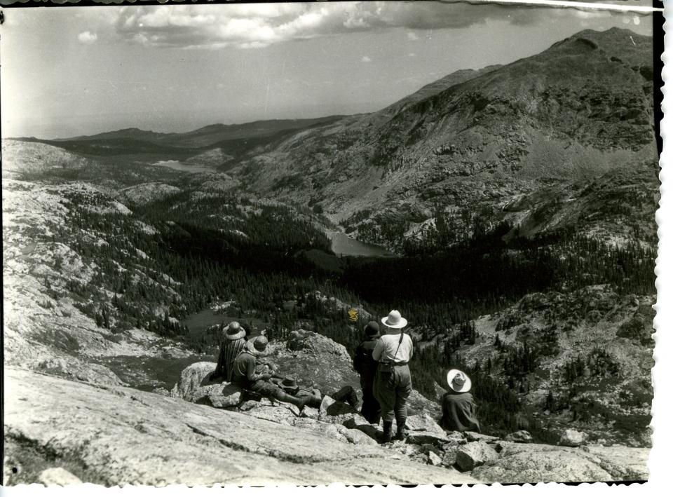 A group of four or five people above timberline look down an alpine valley at a lake in the distance