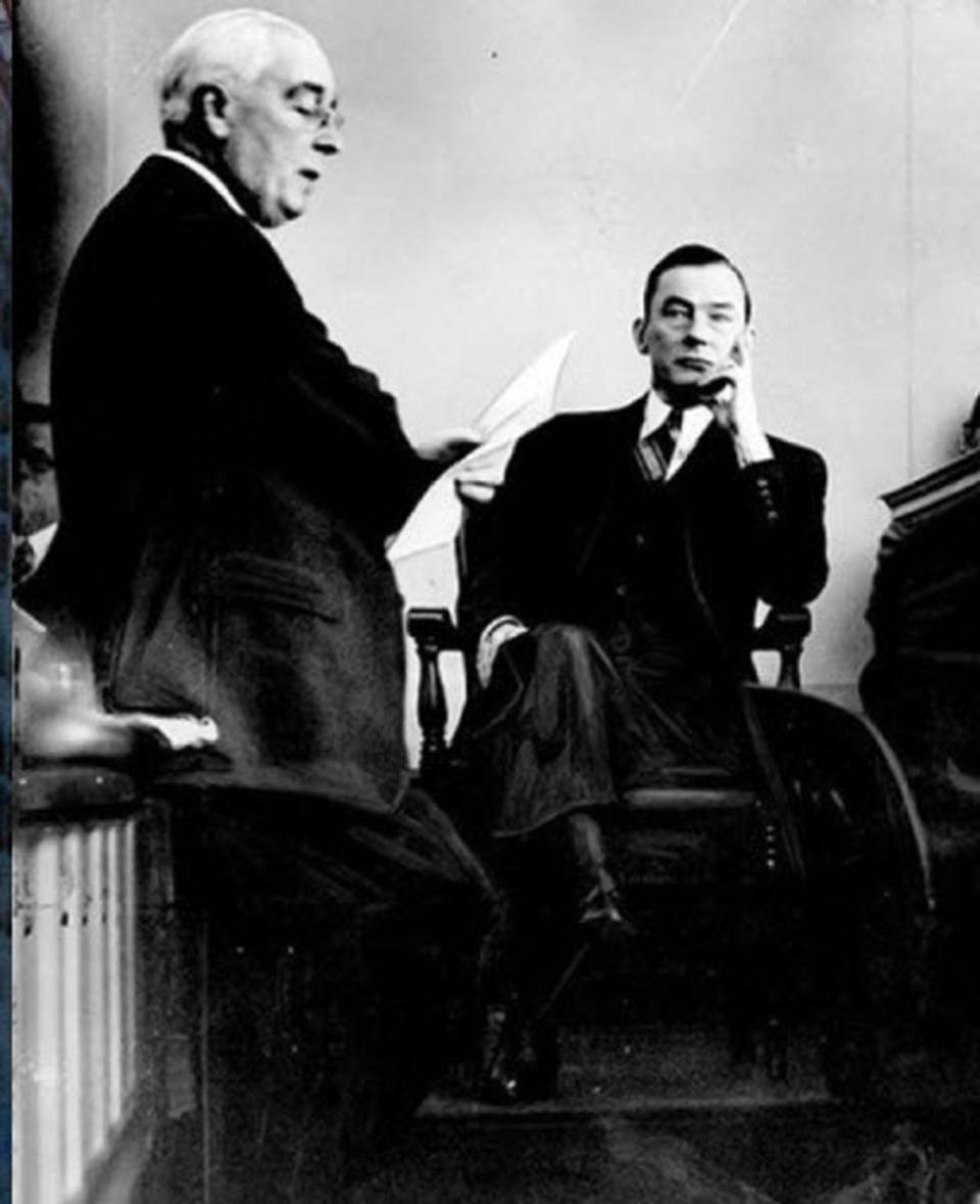 A man in a suit stands and reads from a paper while another man sitting beyond him looks on skeptically appears to read from