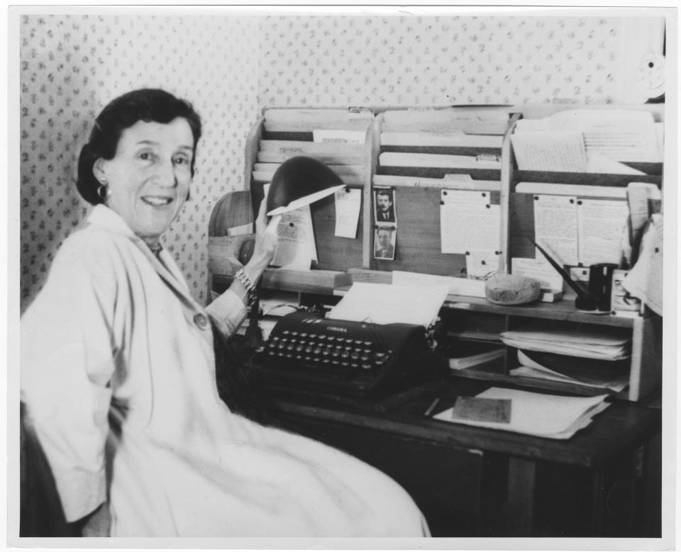 Woman smiles at the camera while sitting at a desk with a typewriter and a lot of file folders and papers.