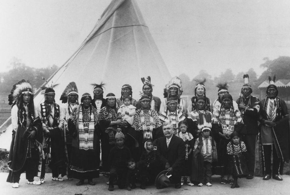 A large group of Northern Arapaho traveled to London and Paris in 1923-24 to appear in live performances staged before showings of the new movie "The Covered Wagon."  Few of the photos show women and children as well as the men; here, the group poses in a park in London. Ed Farlow kneels front and center, Yellow Horse stands at far right and Jack Shavehead stands third from right. The rest are unidentified. American Heritage Center.