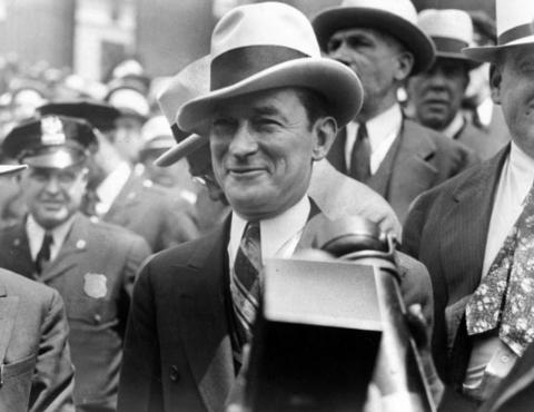 Man in a fedora smiles while posed in front of a large group of men with a policeman looking on