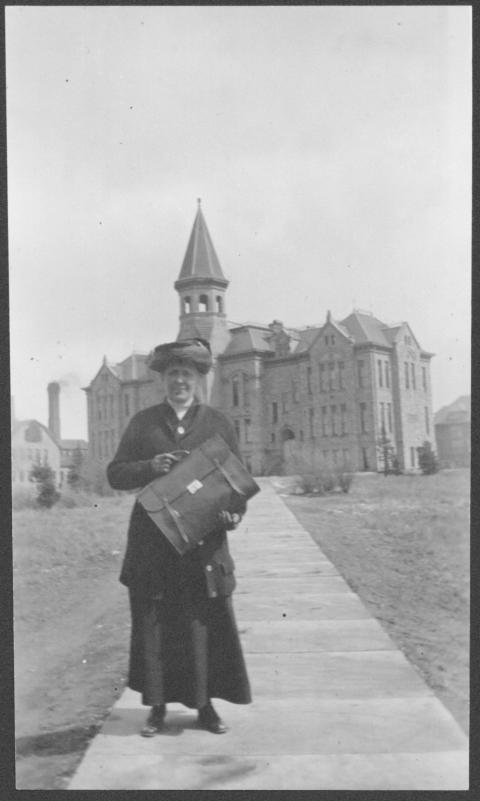 Woman in glasses, long black dress, jacket, hat and gloves posesWhen Grace Raymond Hebard arrived at the University of Wyoming in 1891, Old Main, shown behind her, was the university. Photo File – Hebard, Grace Raymond, University of Wyoming, American Heritage Center. holding a leather briefcase, with an ornate stone building in the background